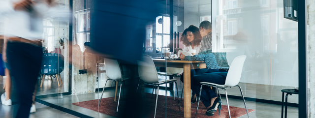  people at a table in a meeting room with an open door and glass walls. Folks are walking by the meeting room.